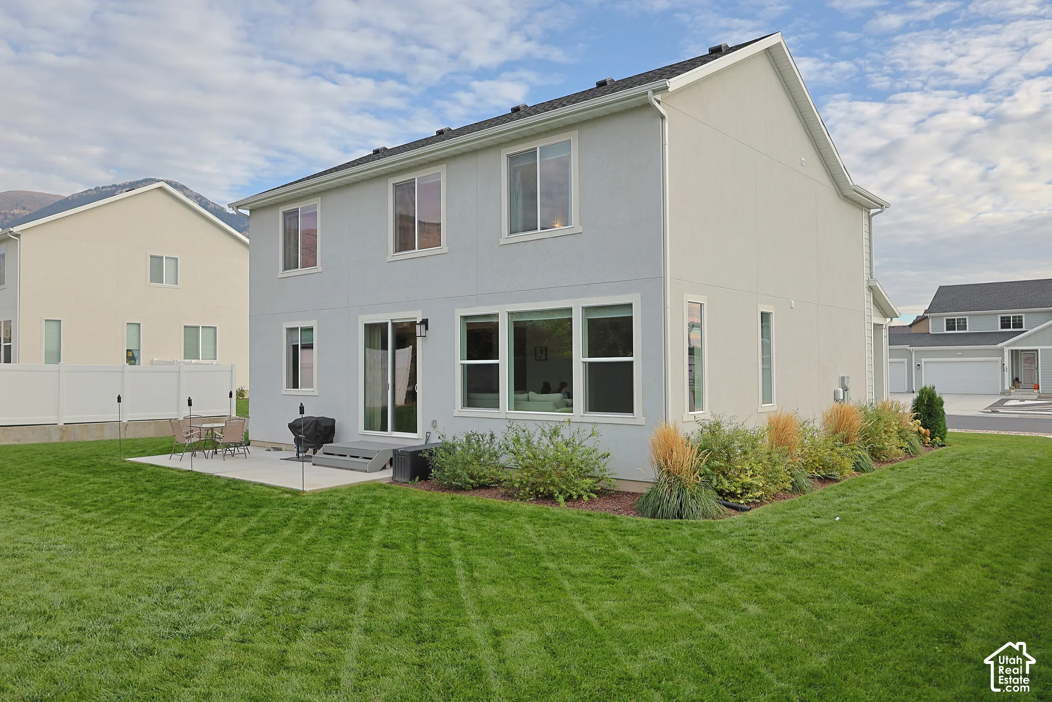 Two-story residence exterior featuring light-toned siding, a concrete patio, large multi-pane windows, sliding glass doors, and a manicured lawn with structured landscaping