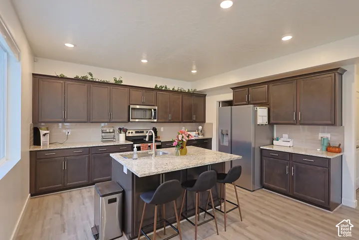 Kitchen featuring dark-finish cabinetry, stone-surface countertops, recessed ceiling lighting, a central island with an integrated sink, and wood-finish flooring