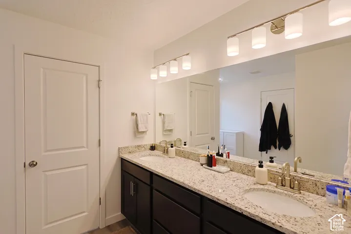 Dual-sink vanity featuring stone-finish countertops, dark cabinetry, polished metal fixtures, and full-width wall-mounted mirrors with multi-bulb light bar illumination