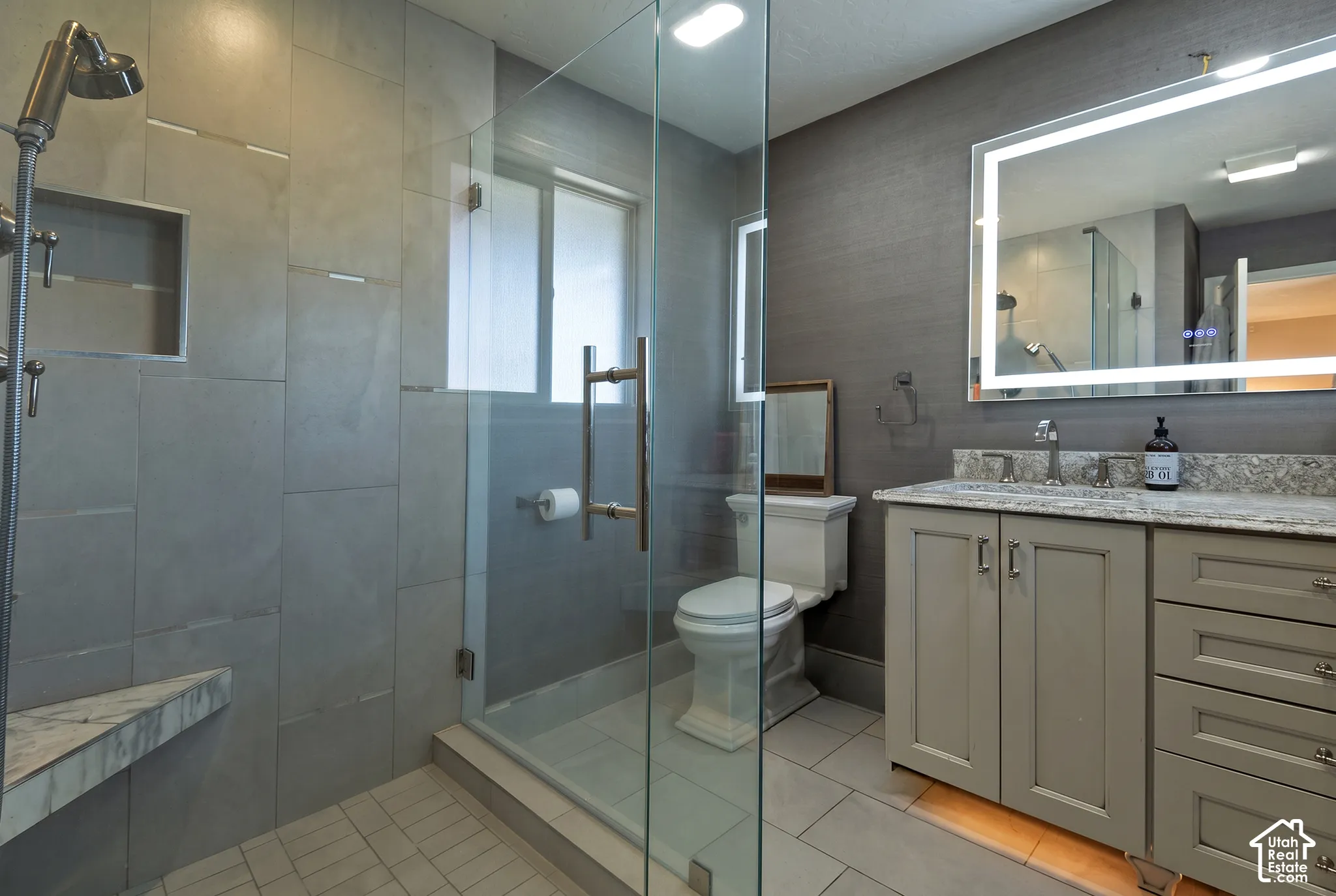 Walk-in shower with large-format tile surround, stone-finish countertop, built-in cabinetry with drawer storage, and a wall-mounted LED mirror