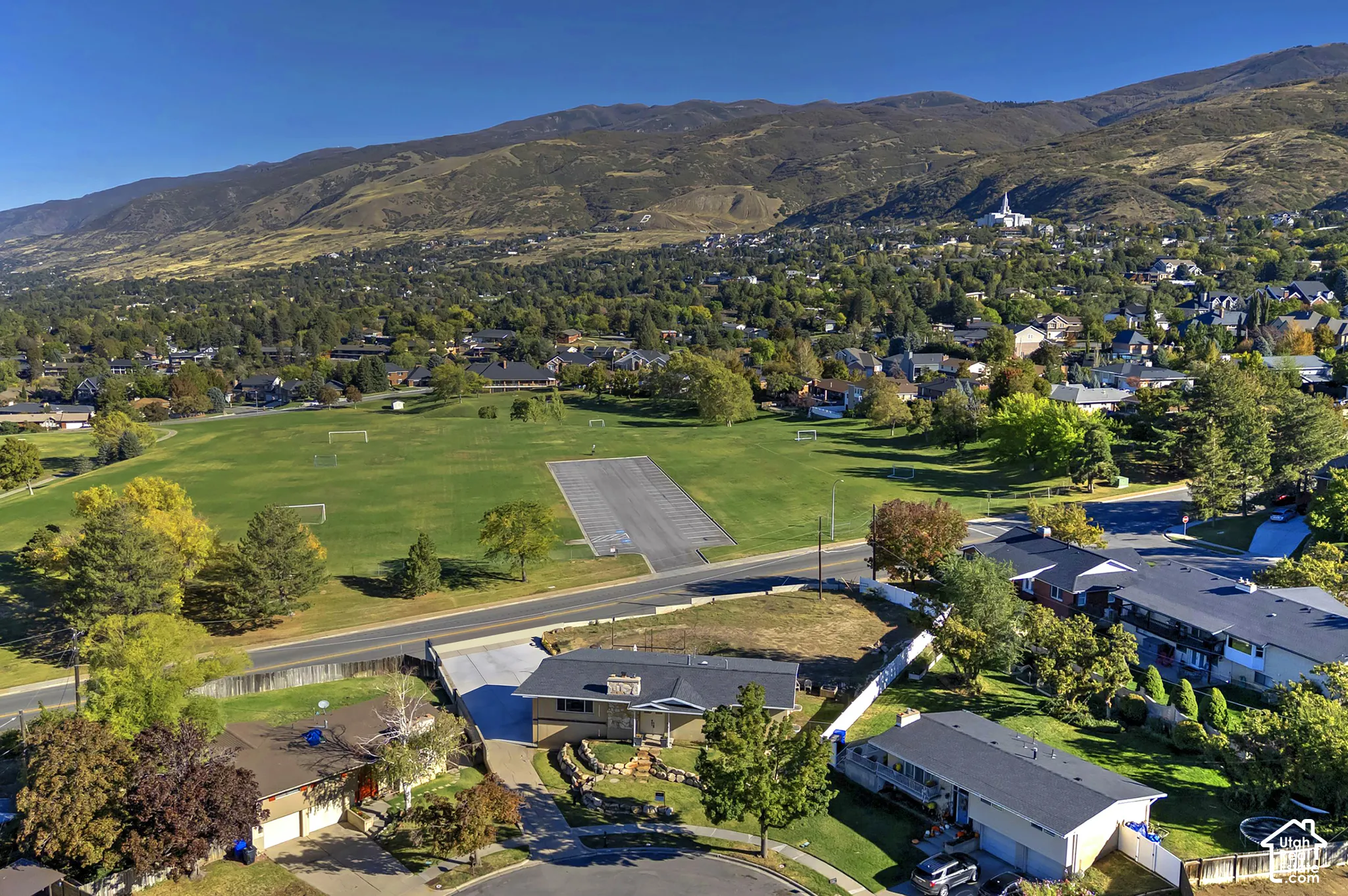 Aerial perspective of a single-story residence featuring a stone-clad chimney, a concrete driveway, tiered stone retaining walls, and mountain scenery