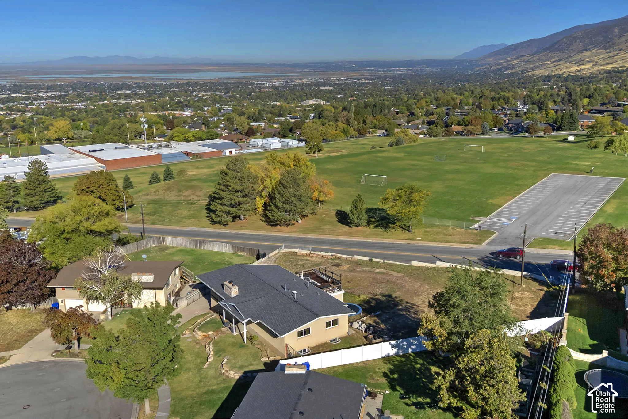 Aerial perspective of a multi-level residence featuring a pitched asphalt shingle roof, exterior stone chimney, private fenced yard, and mountain backdrop