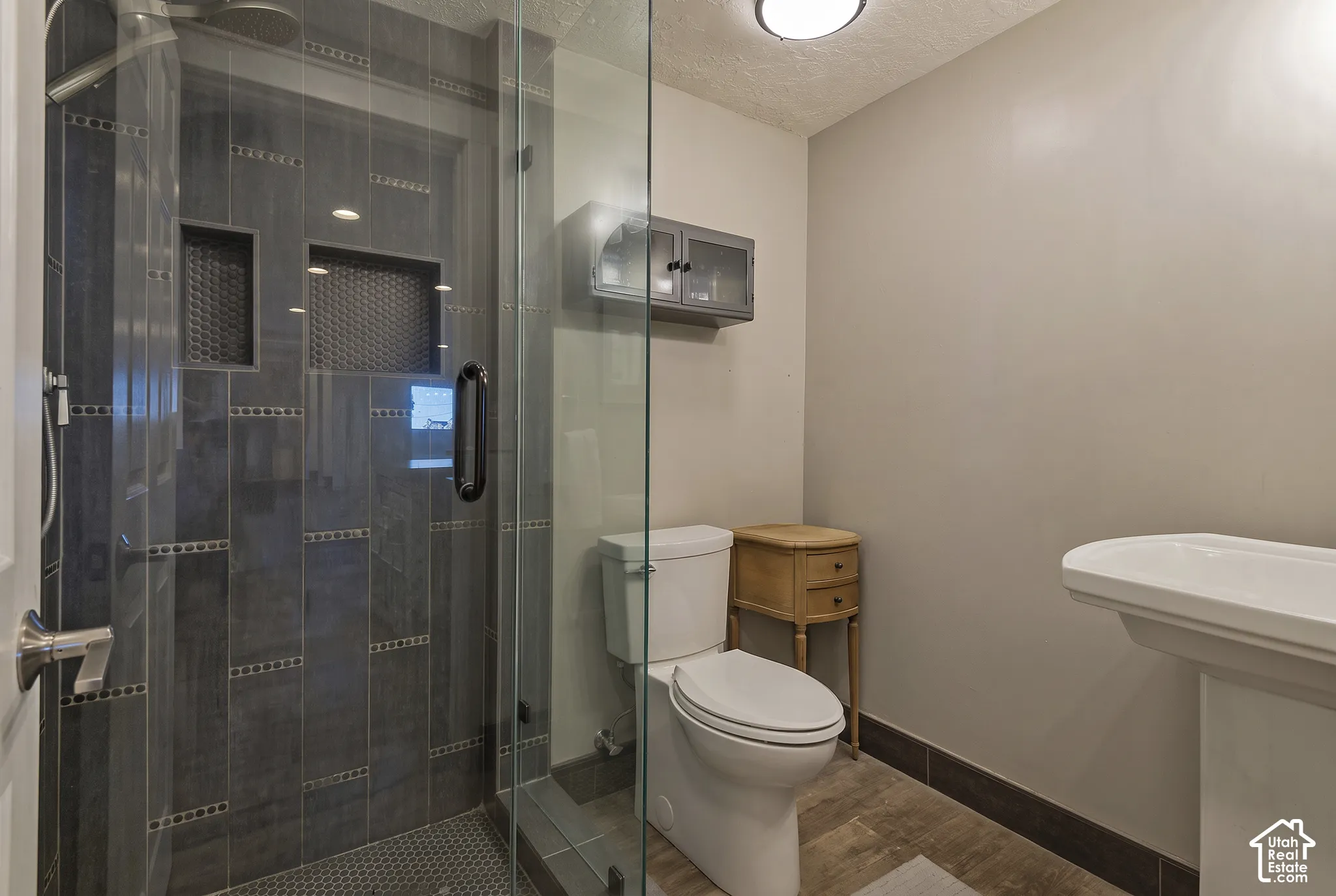Walk-in shower with floor-to-ceiling tile, inset wall niches, glass door enclosure, pedestal sink, and wood-finish flooring