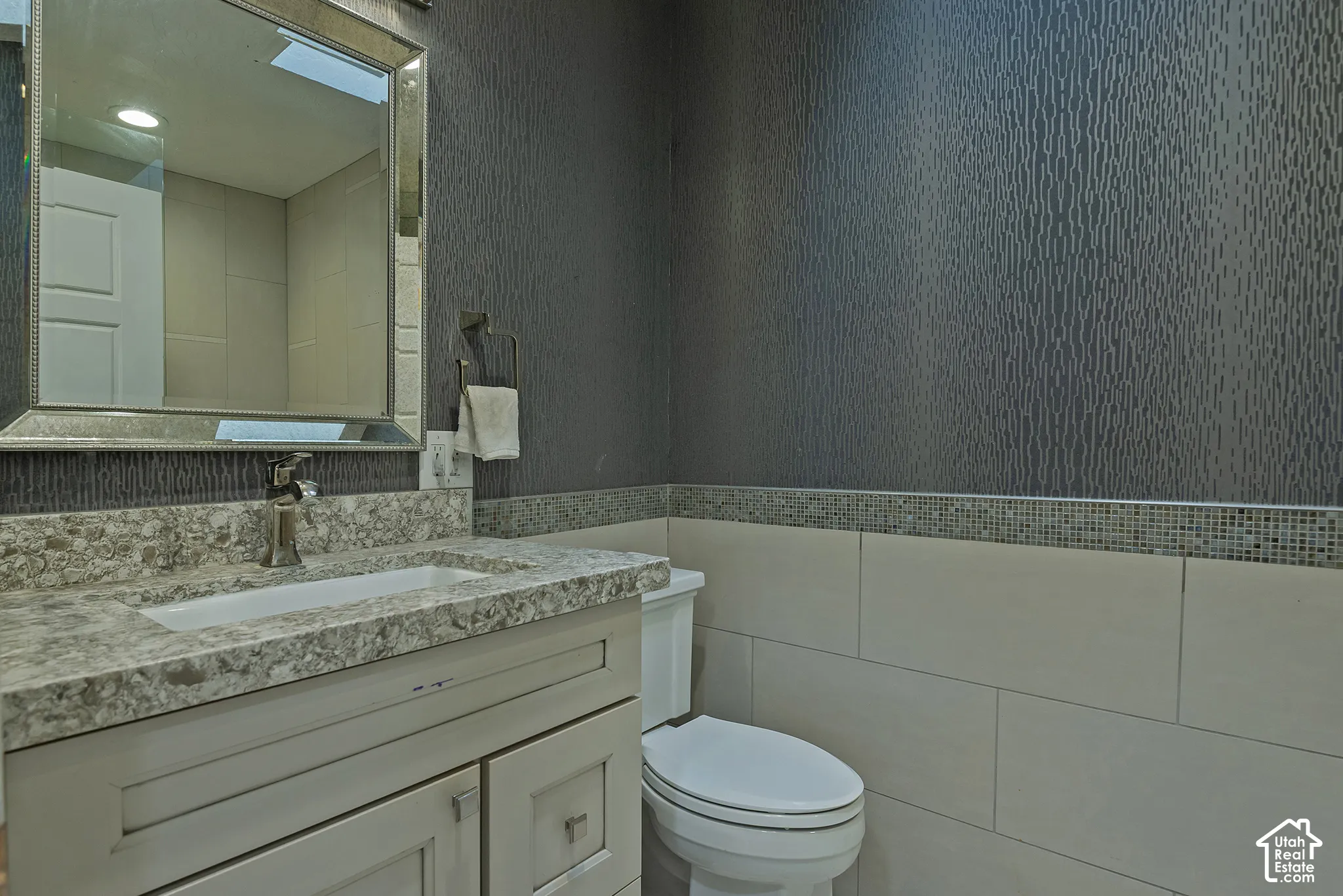 Vanity with stone-finish countertop and integrated sink, framed wall mirror, textured wallpaper, mosaic tile accent border, and large-format wall tiling