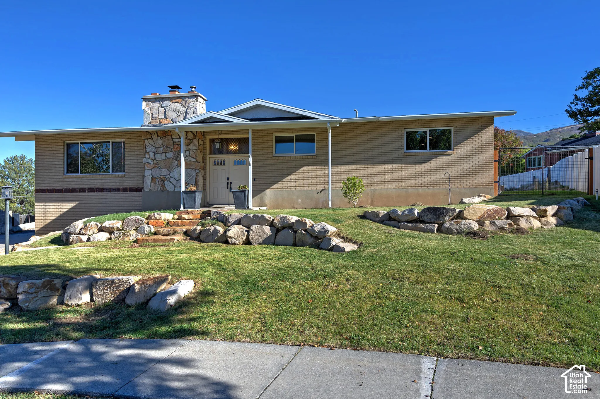 Single-story brick exterior featuring a prominent stone chimney, terraced landscaping with stacked-stone retaining walls, and double-entry doors with transom windows