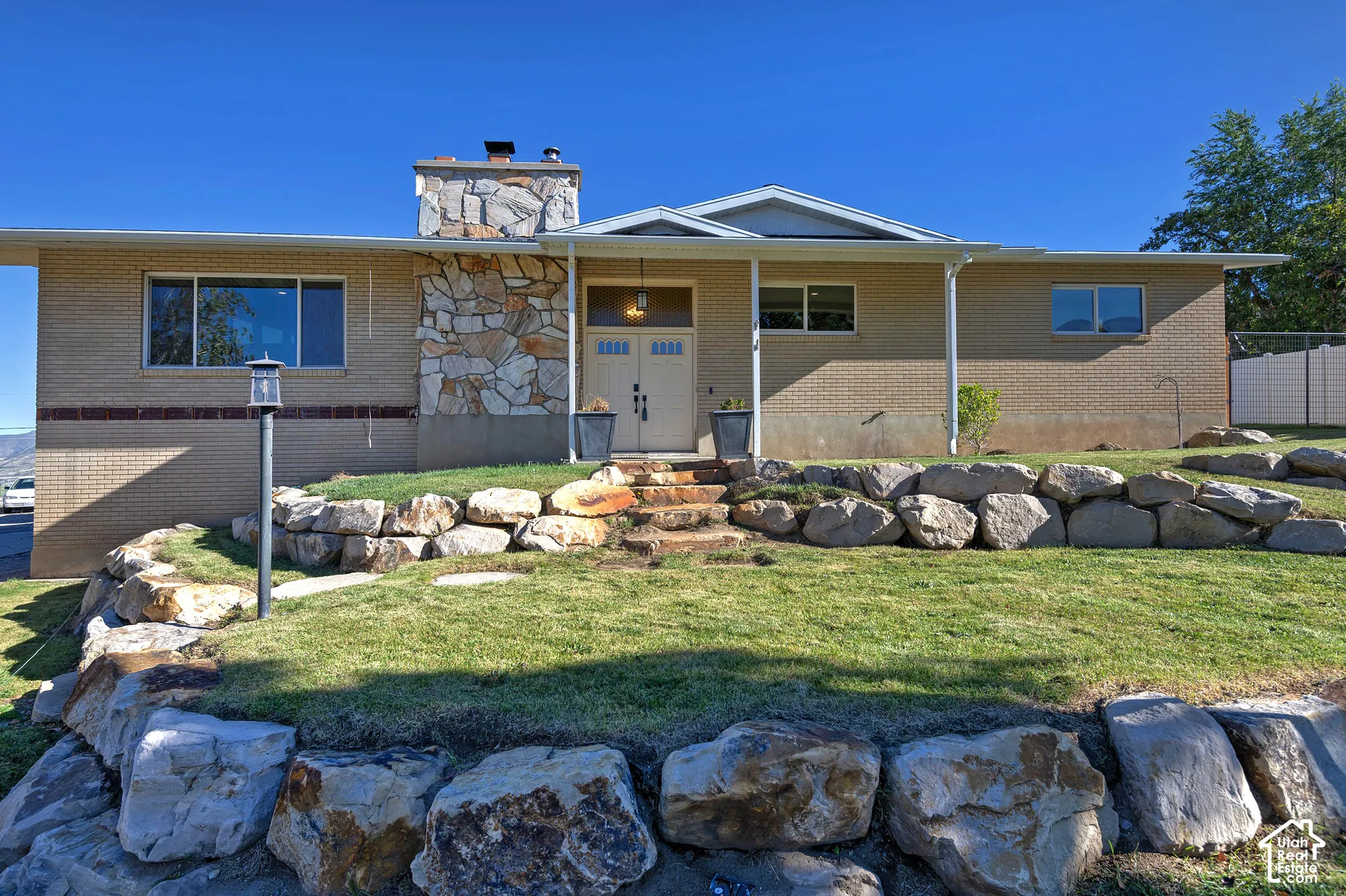 Single-level brick exterior featuring a multi-tonal stone chimney, stacked stone retaining walls, and a dual-entry front door with glass transom