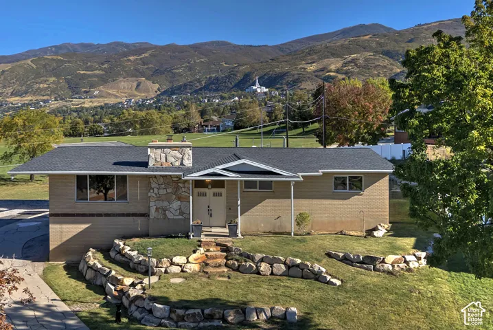 Single-story residence featuring a brick exterior, stone chimney detailing, dual-entry front doors, and a terraced rock-retaining wall, set against a backdrop of mountain scenery