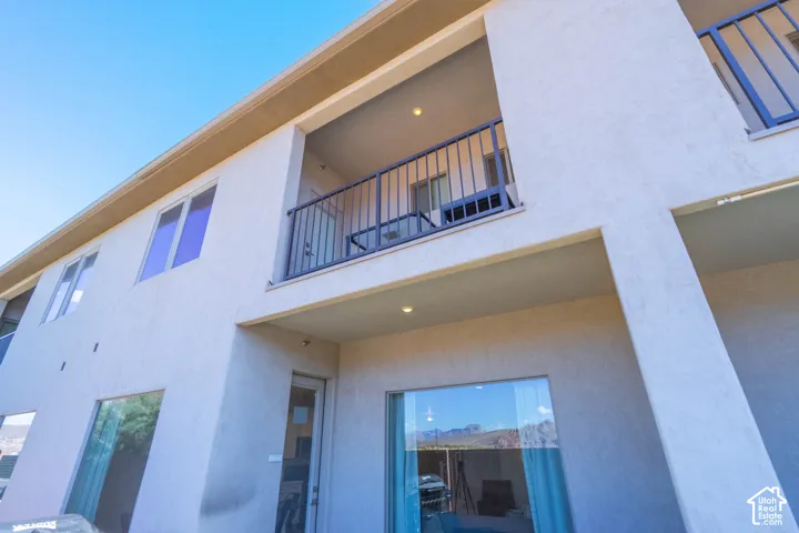Exterior stucco facade featuring a second-level balcony, dark metal railing, recessed ceiling lighting, and large picture windows