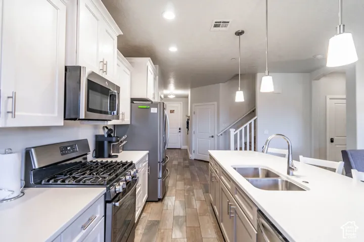 Kitchen featuring white cabinetry, wood-finish flooring, a quartz-topped center island with an undermount dual-basin sink, stainless steel appliances, and pendant lighting