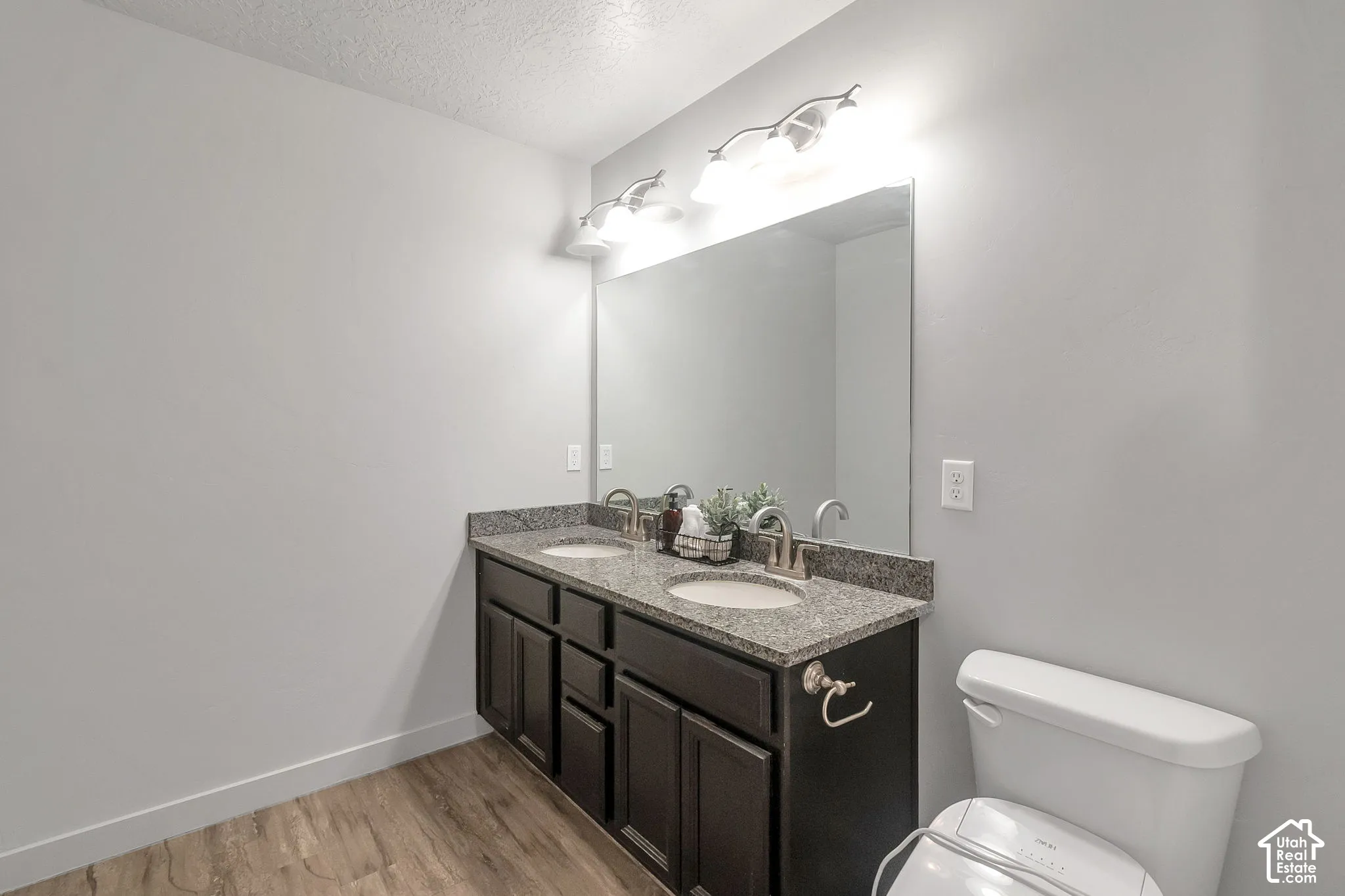 Double vanity with stone-finish countertop, dual drop-in sinks, dark cabinetry, and full-width mirror with three-bulb vanity lighting