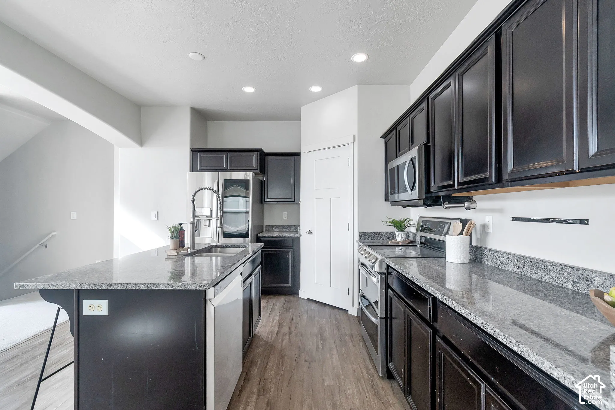Kitchen featuring dark-finish cabinetry, stone-surface countertops, stainless steel appliances, a center island with a dual-basin sink, and wood-finish flooring