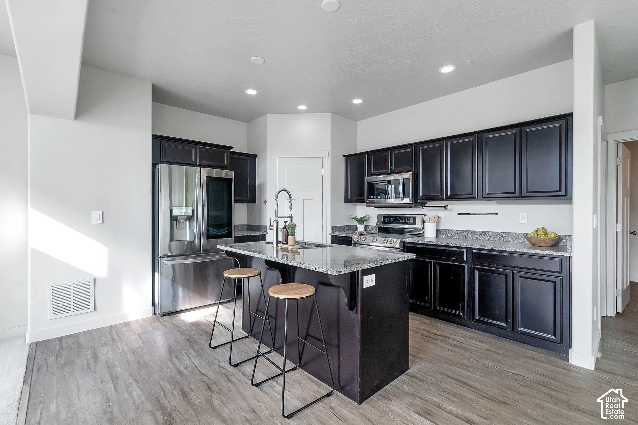 Kitchen featuring dark cabinetry, stone-finish countertops, stainless steel appliances, central island with integrated sink, and wood-finish flooring