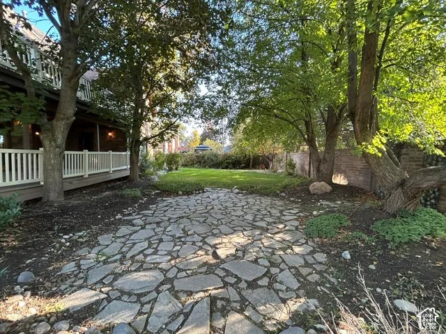 Backyard middle area featuring an irregular stone patio, mature trees, a manicured lawn, a white wooden deck railing, and a masonry perimeter wall