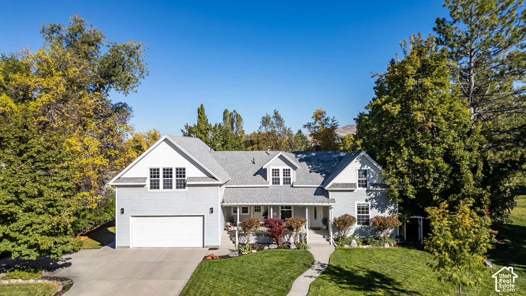 Multi-level residence featuring light-toned horizontal siding, a covered front porch, a two-car garage, and a manicured lawn surrounded by mature trees