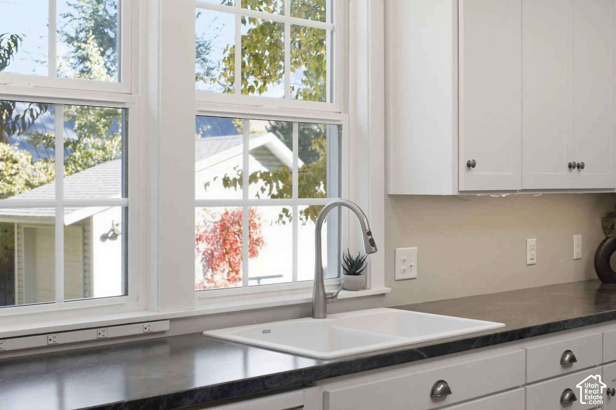 Kitchen featuring white shaker-style cabinetry, dark stone-finish countertops, a dual-basin white sink with a gooseneck faucet, and white mullioned windows