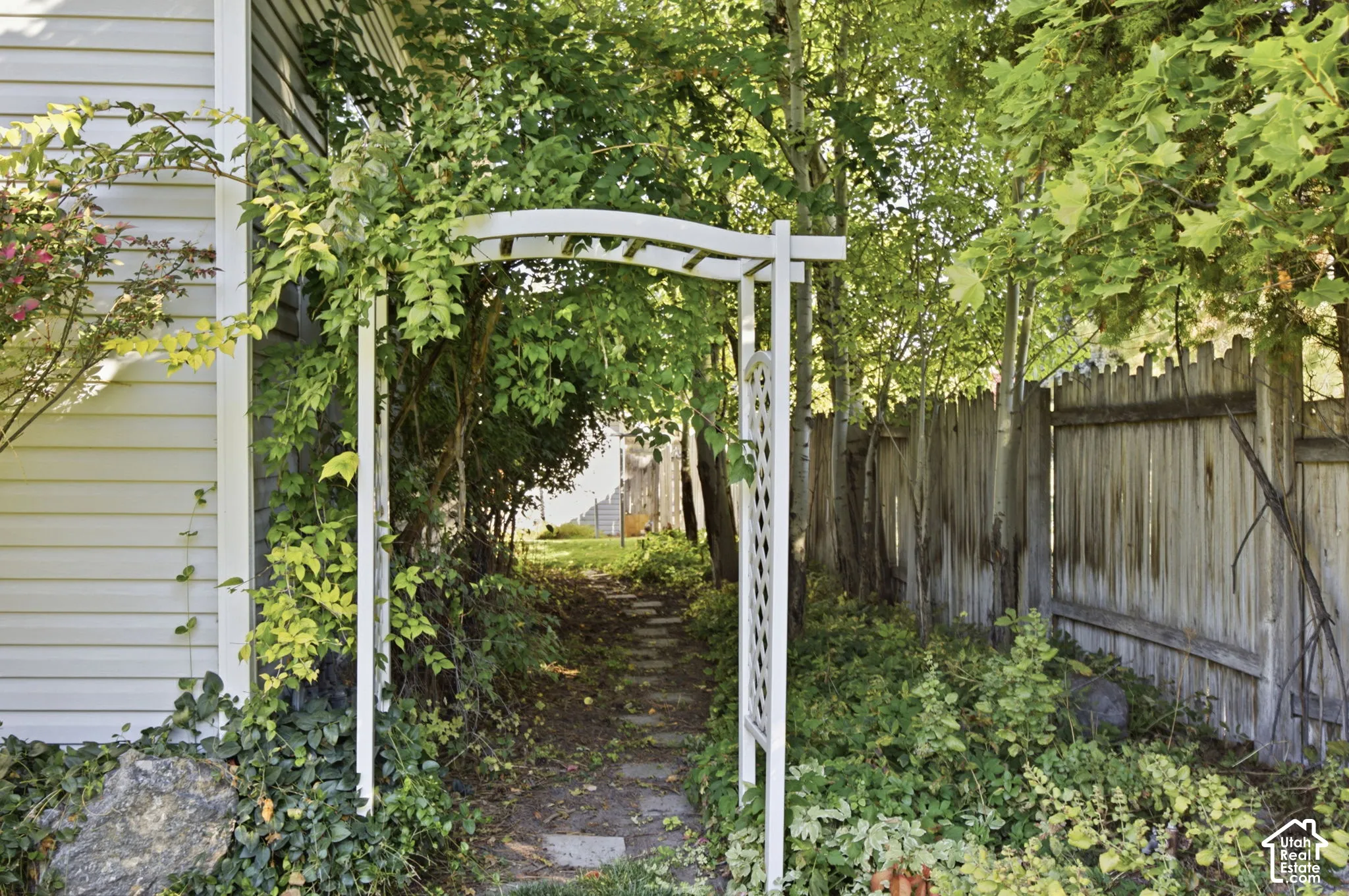 White garden arbor with lattice detailing, stone paver walkway, mature foliage, and wood privacy fencing