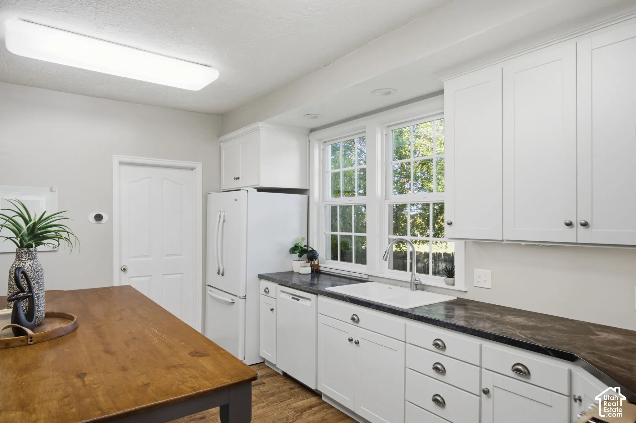 Kitchen featuring white shaker-style cabinetry, dark stone-finish countertops, a white undermount farmhouse sink, and wood-finish flooring