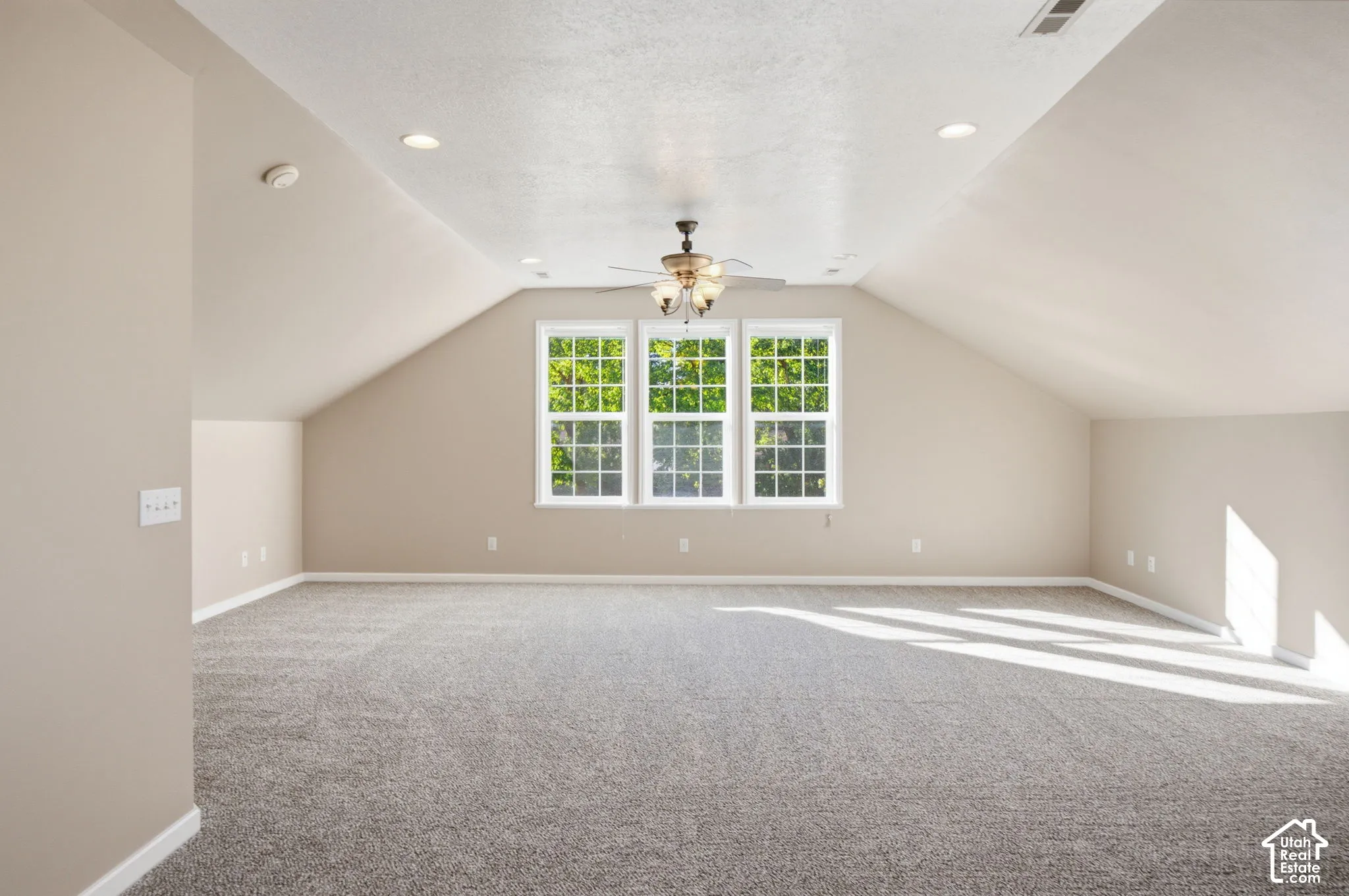 Spacious room featuring vaulted ceilings, neutral wall-to-wall carpeting, recessed lighting, a ceiling fan with integrated light fixture, and a triple-pane window configuration