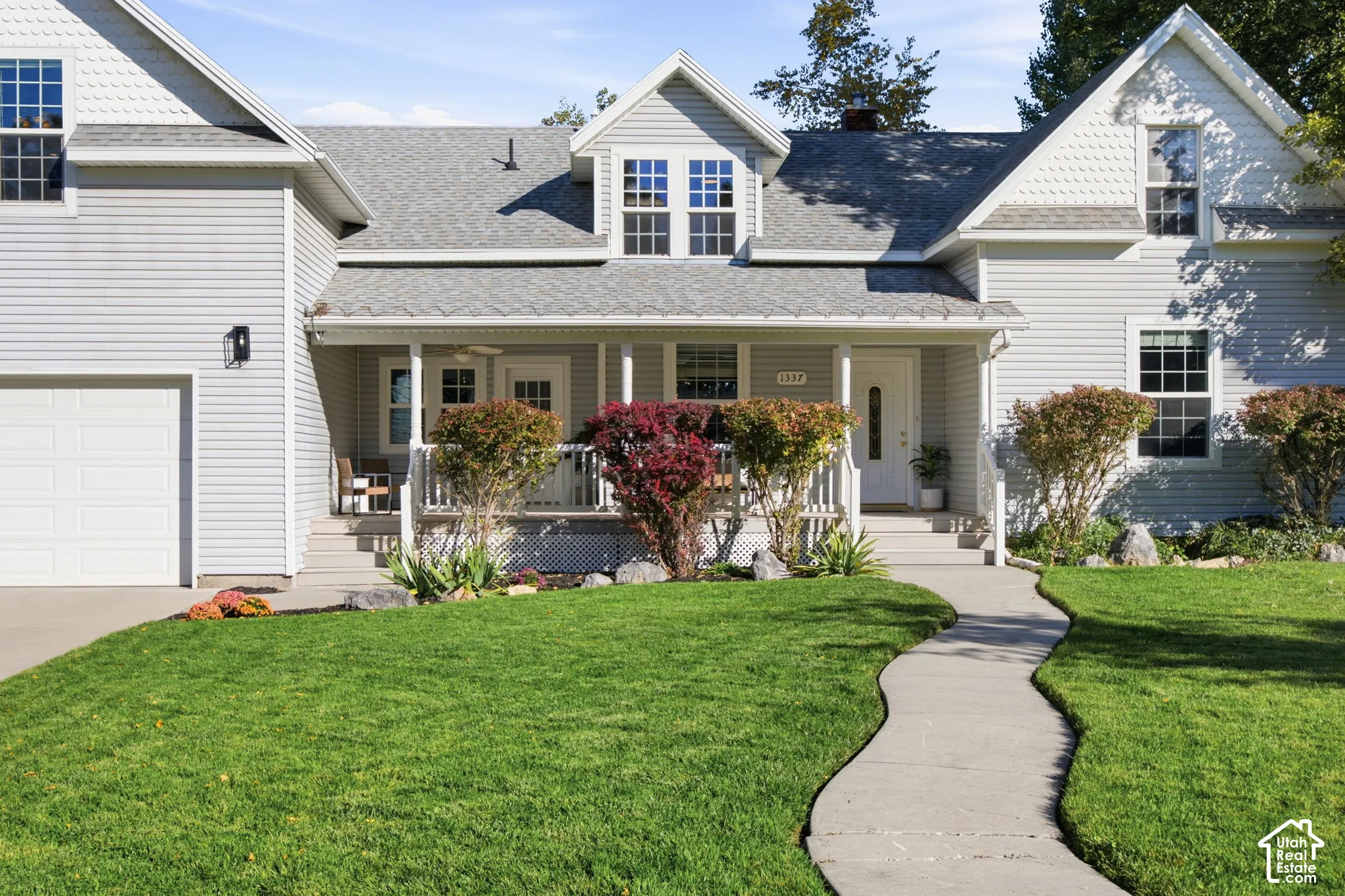 Two-story residence featuring light gray siding, gabled rooflines with decorative shingles, a covered front porch with white railing, and a paved walkway leading to the entrance
