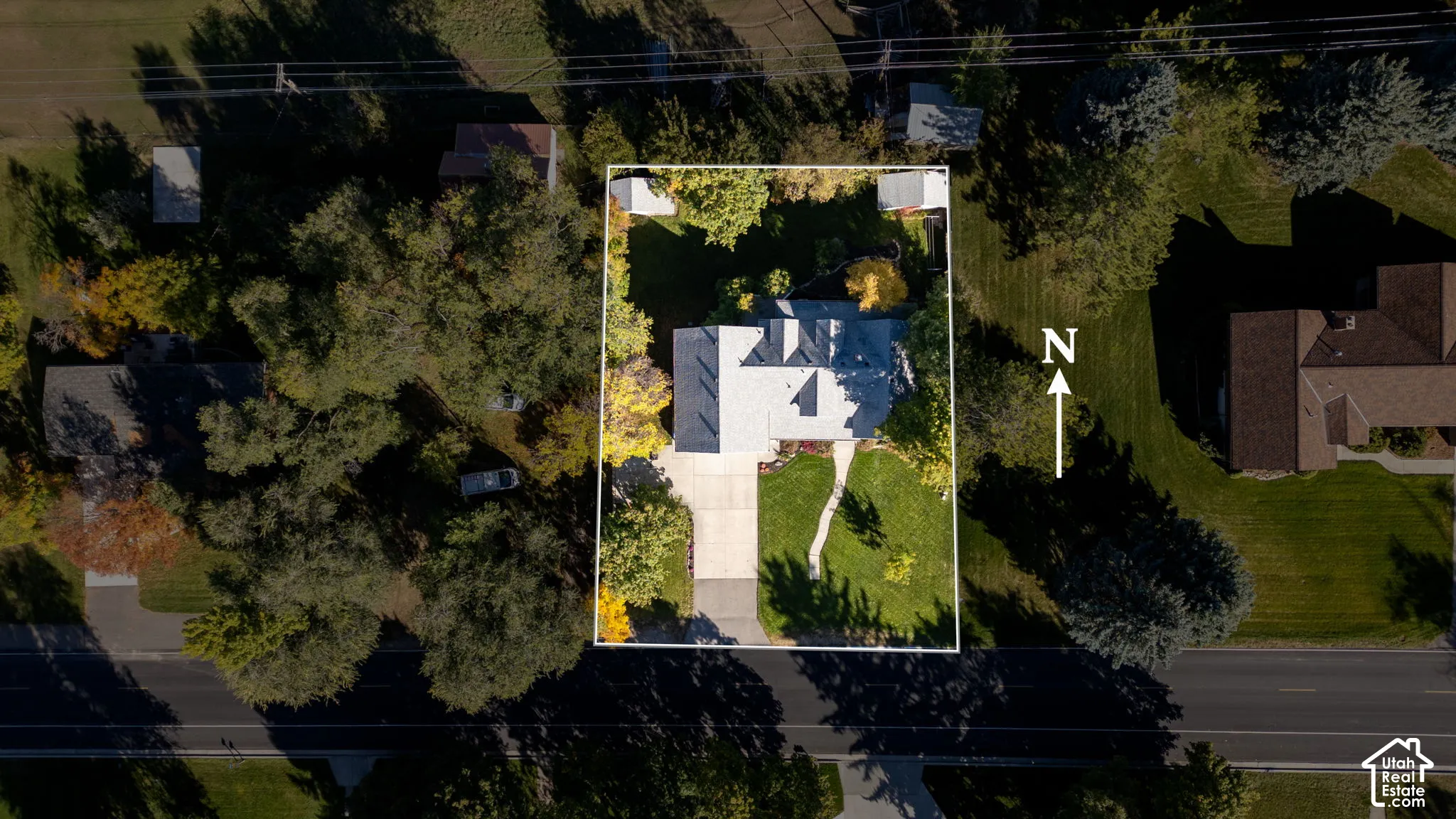 Aerial perspective of a property featuring a rectangular lot, mature perimeter landscaping, concrete driveway, stone-paved walkway, and two detached storage structures
