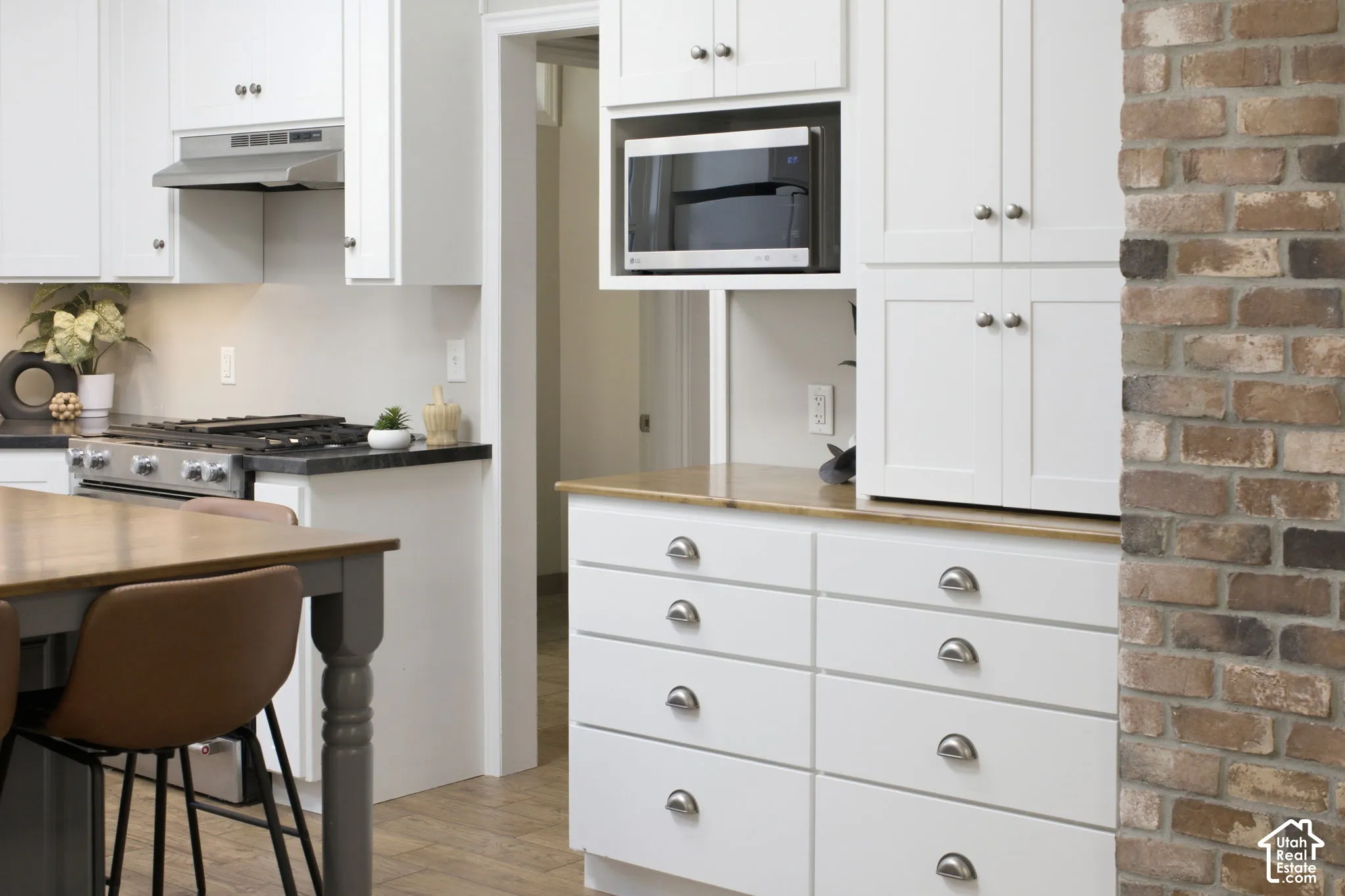 Kitchen featuring white shaker-style cabinetry, wood-finish countertops, exposed brick accent wall, stainless steel range with range hood, and integrated built-in microwave