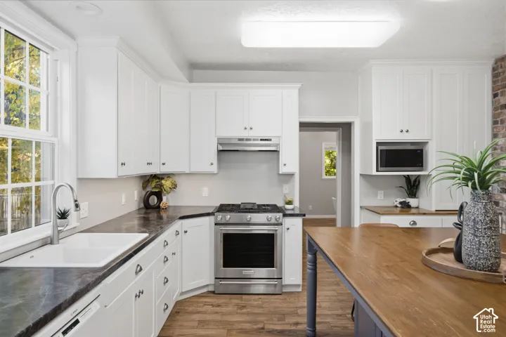 Kitchen featuring white shaker-style cabinetry, dark stone-finish countertops, wood-finish flooring, stainless steel gas range, and a dual-basin undermount sink
