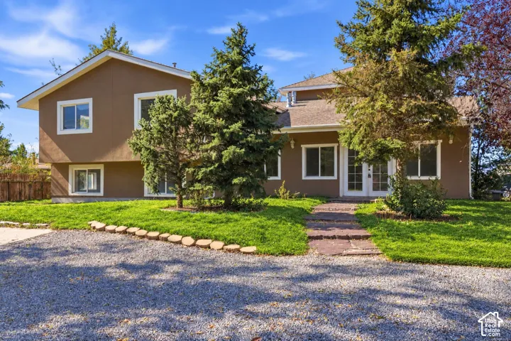 Two-story exterior with stucco finish, stone-tiled walkway, gravel driveway, and mature conifer trees