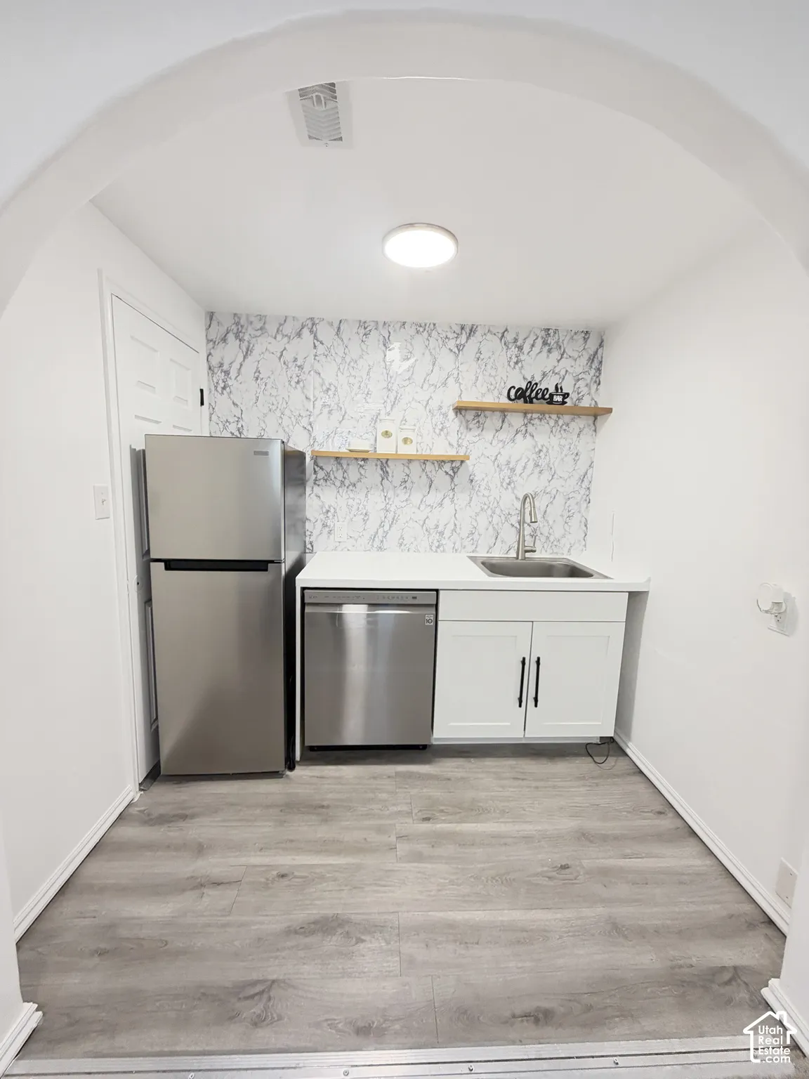 Kitchenette featuring a stainless steel refrigerator and dishwasher, white cabinetry with matte black hardware, stone-patterned backsplash, wood-finish flooring, and floating wooden shelving