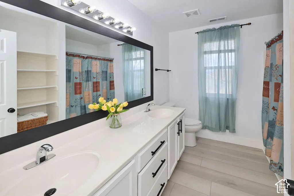 Dual-sink vanity with quartz countertop, expansive wall-to-wall mirror, white cabinetry with matte black hardware, built-in shelving, and wood-finish flooring
