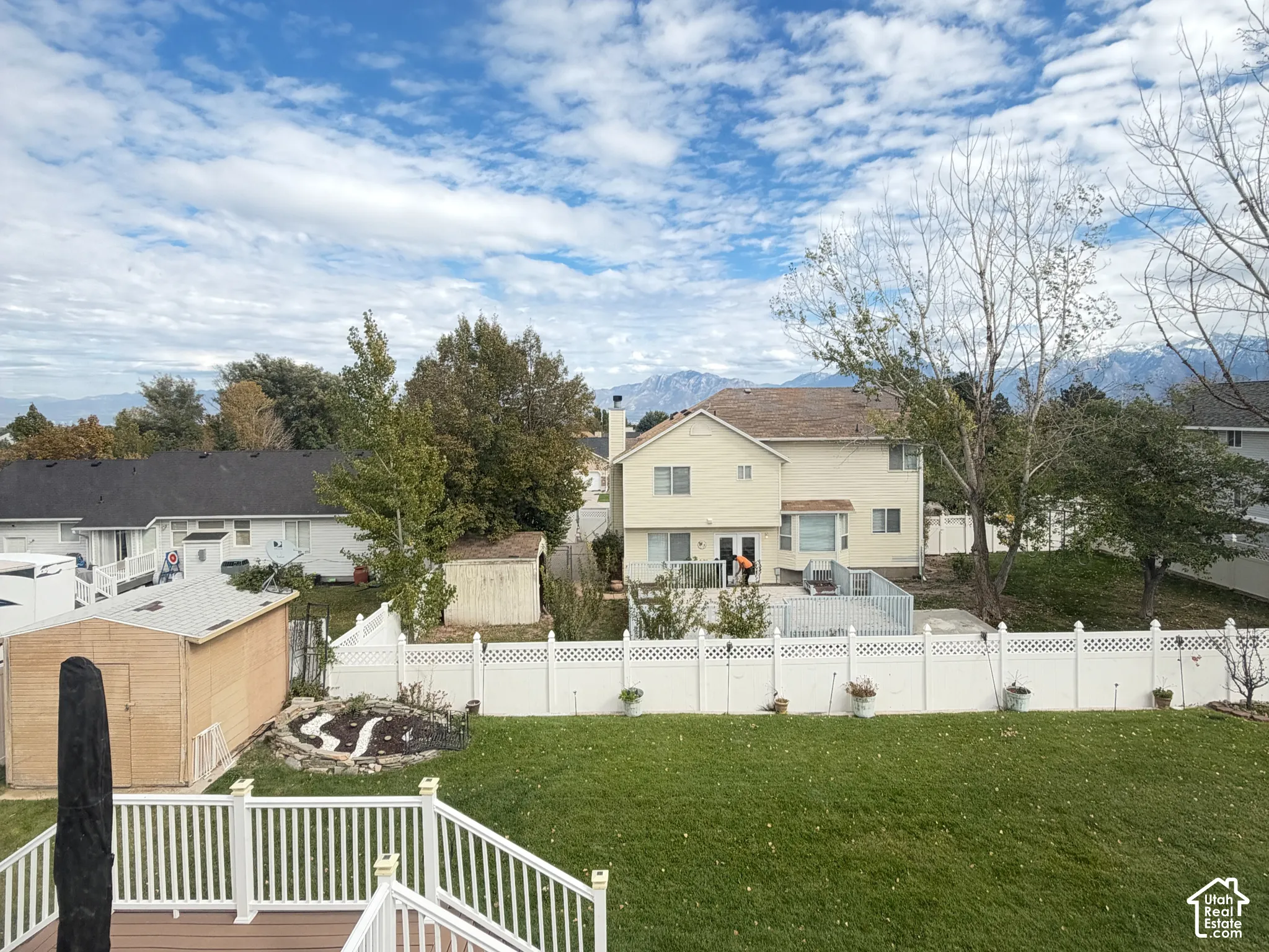 Elevated deck with white vinyl railing, stone-paver patio, expansive lawn, white vinyl fencing, and mountain backdrop