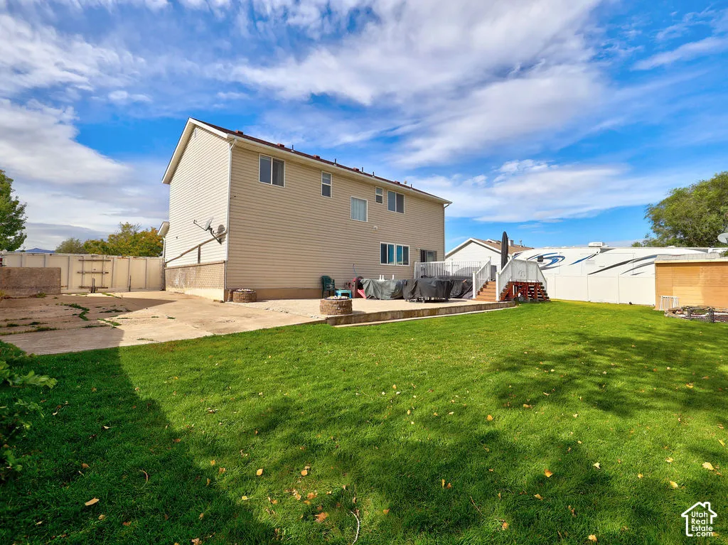 Expansive backyard featuring a manicured lawn, concrete patio, built-in fire pit, elevated wooden deck, and full-perimeter vinyl fencing