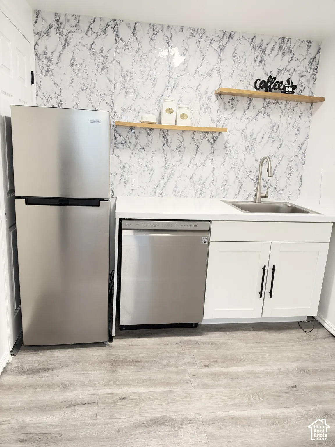 Kitchenette featuring stone-patterned backsplash, white cabinetry with matte black hardware, integrated stainless steel dishwasher, stainless steel refrigerator, and wood-finish flooring