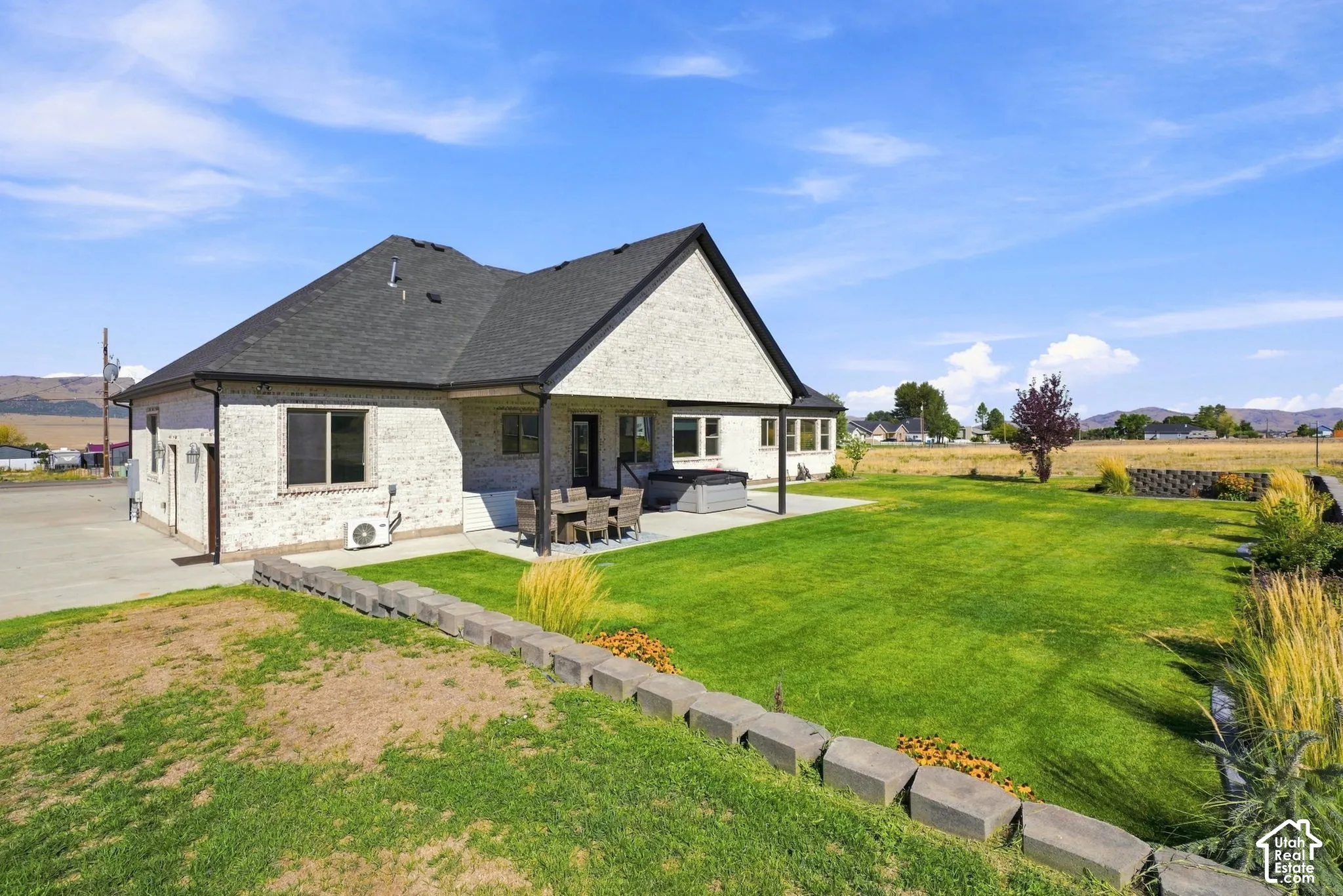 Brick exterior residence featuring a covered rear patio, expansive lawn, stone block retaining wall, and mountain backdrop