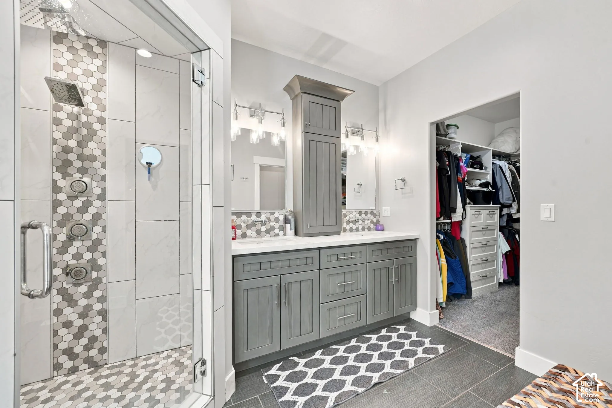 Dual-vanity bathroom featuring a custom grey cabinetry unit with a built-in tower, multi-head walk-in shower with decorative tile inlay, and built-in walk-in closet shelving