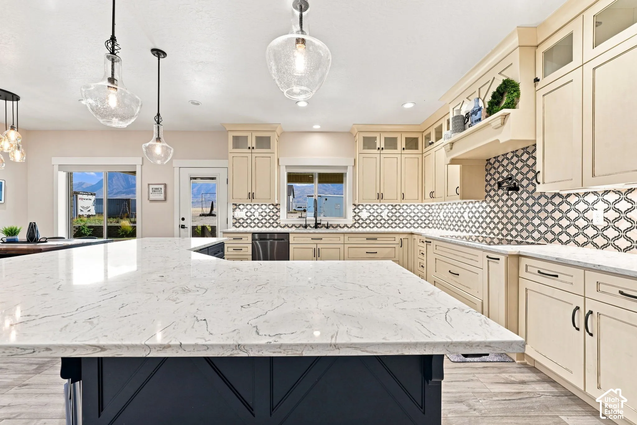 Kitchen featuring stone-finish countertops, a contrasting dark-finish island base, patterned tile backsplash, custom cabinetry with glass-panel upper doors, and glass-pendant lighting