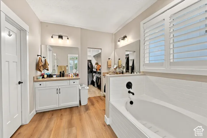 Bathroom featuring wood-finish flooring, a soaking tub with subway tile surround, white cabinetry with a solid surface countertop, wall-mounted matte black plumbing fixtures, and plantation shutters