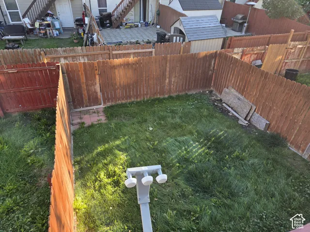 Fenced backyard featuring a grass lawn, hardscaped paver patio area, and mounted security lighting