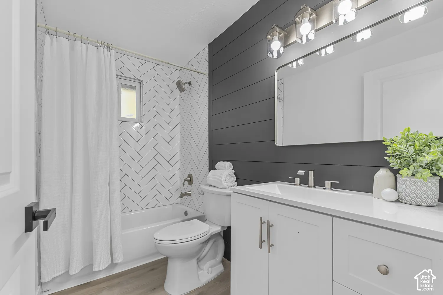 Bathroom featuring a herringbone tile tub surround, charcoal horizontal shiplap wall accent, white cabinetry with a surface-mounted sink, wood-finish flooring, and a multi-bulb vanity light fixture