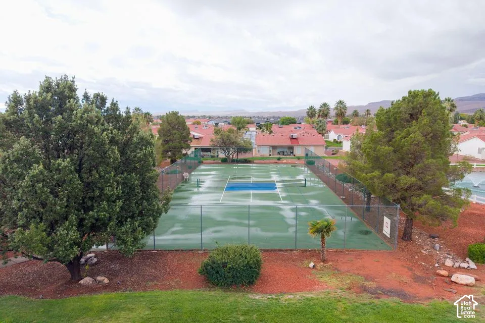 Tennis court enclosed by chain-link fencing, situated within a landscape of mature trees, shrubs, and a grass lawn