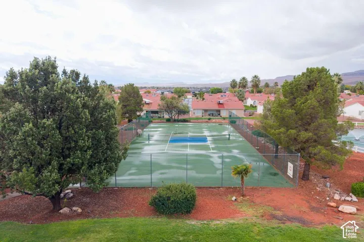 Tennis court enclosed by chain-link fencing, situated within a landscape of mature trees, shrubs, and a grass lawn
