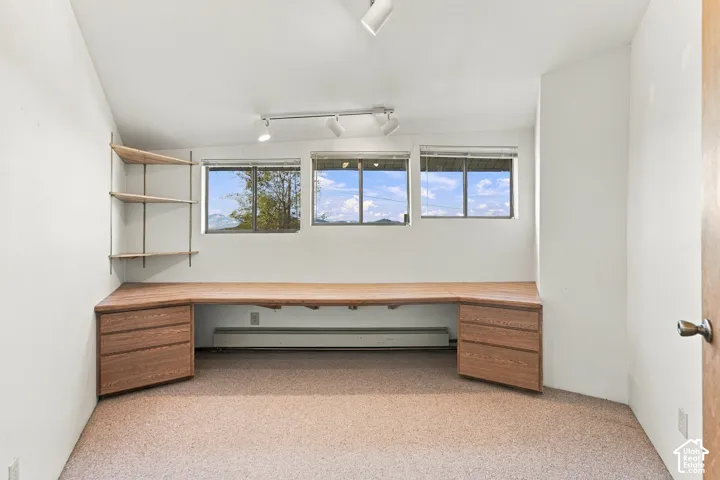 Built-in wood-finish desk with integrated cabinetry and drawers, three large windows, adjustable wall-mounted shelving, and multi-directional track lighting