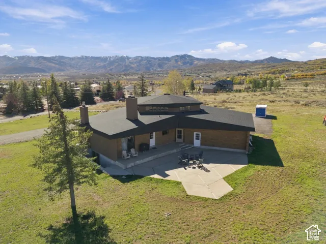 Multi-level property exterior featuring brick masonry, a shingle roof, dual exterior chimneys, and an expansive concrete patio situated against a mountain backdrop