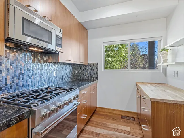 Kitchen featuring wood-finish cabinetry, stone countertops, a stainless steel gas range with a matching microwave, a mosaic tile backsplash, and wood-finish flooring