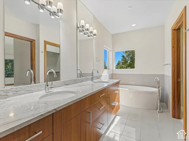 Dual-vanity cabinet featuring wood-finish cabinetry, stone countertop with integrated sinks, polished chrome fixtures, and a soaking tub with a floor-mounted faucet