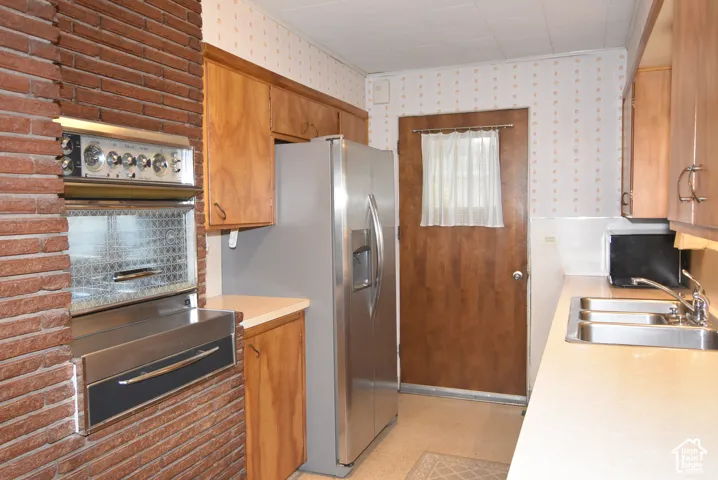 Kitchen featuring a brick chimney wall, built-in dual oven, stainless steel side-by-side refrigerator, ample wood cabinetry, and a triple-basin stainless steel sink