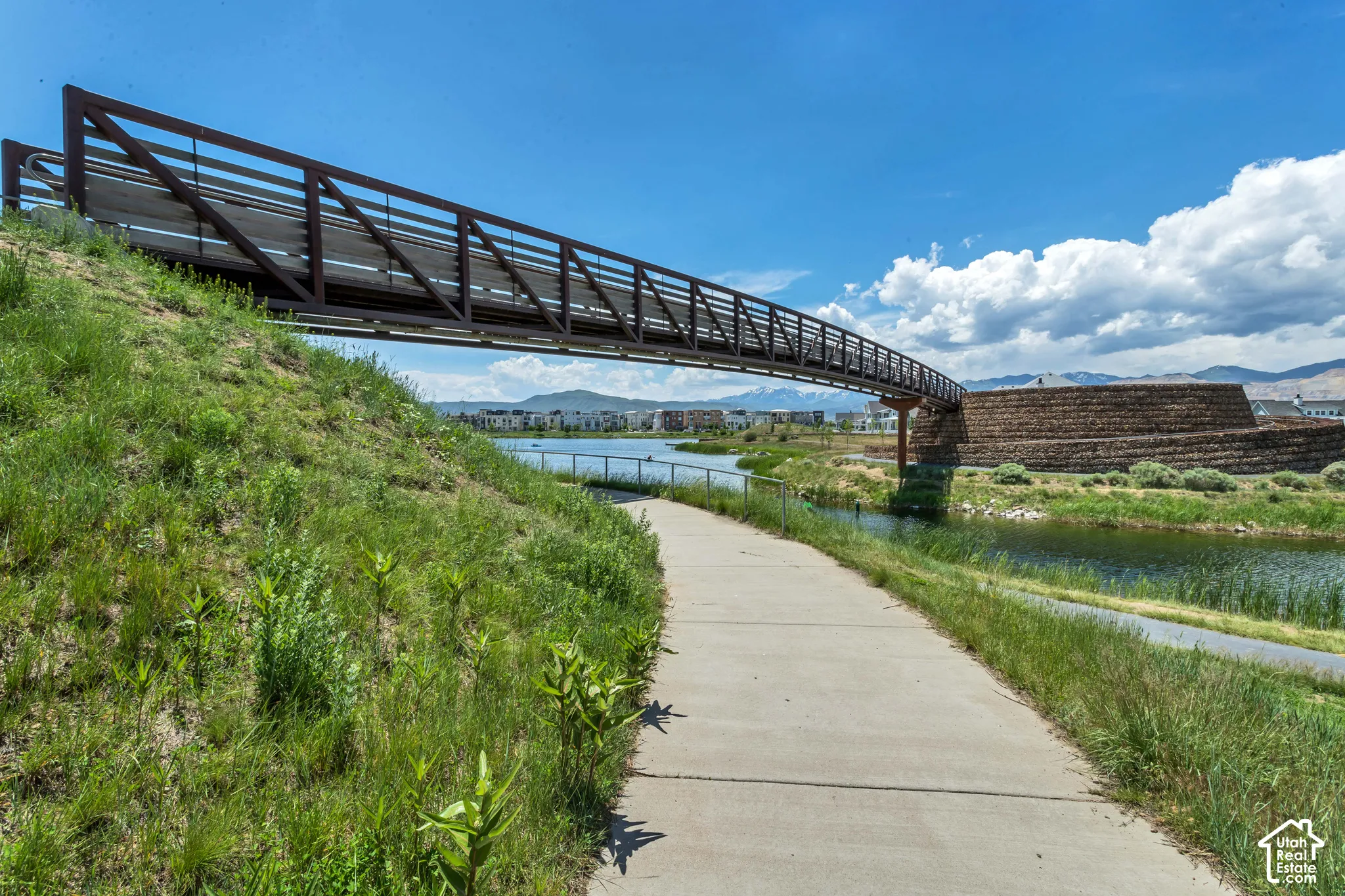 Pedestrian bridge featuring metal railings, concrete path, pond, and stone-retaining wall structures set against a mountain backdrop