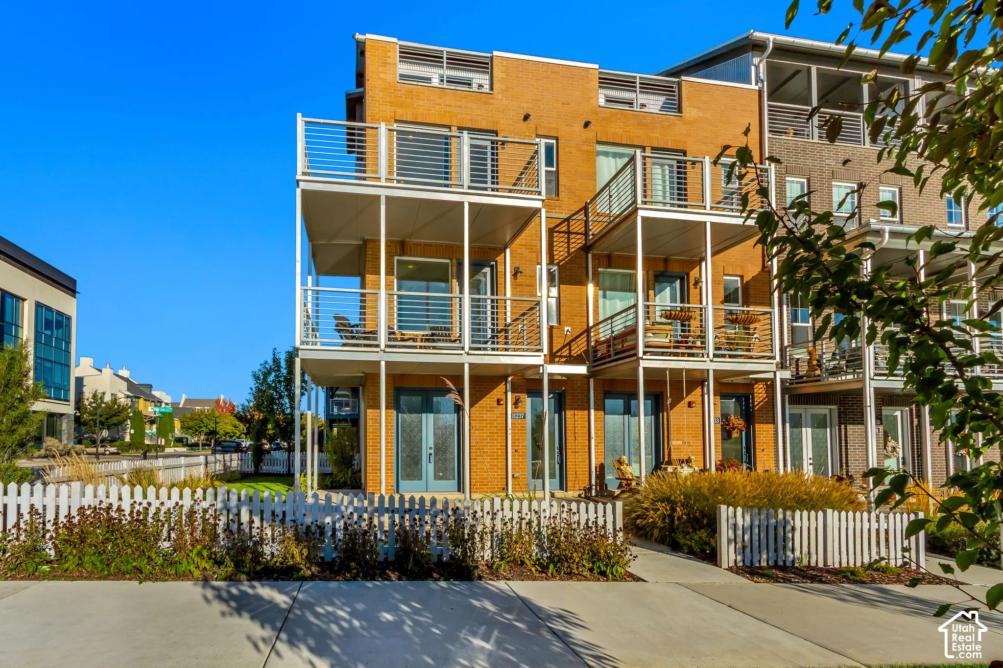 Multi-level brick exterior featuring stacked balconies with metal railings, glass-paneled double doors, and a perimeter picket fence