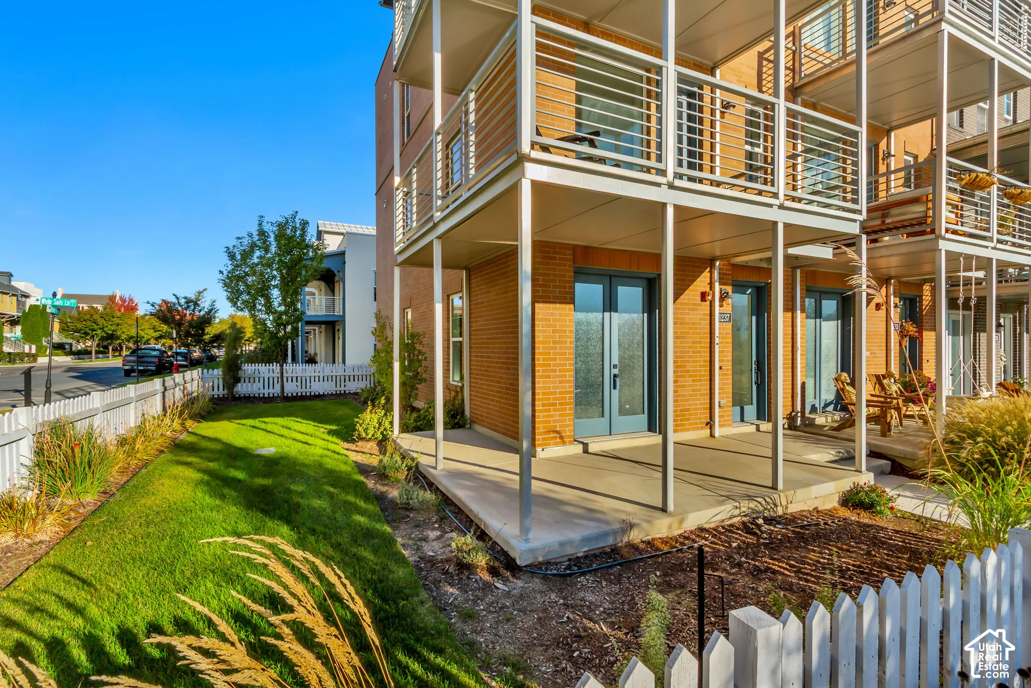 Multi-story brick exterior featuring private balconies with metal railing, concrete ground-level patio, double-door entry, and white picket fencing