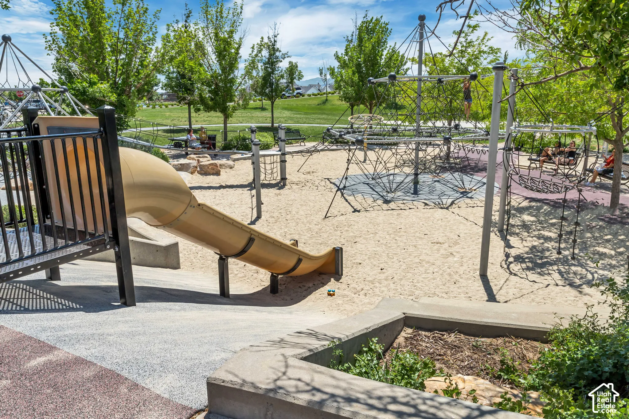 Community playground area featuring structured climbing apparatus, a tube slide, sand surfacing, and mature trees