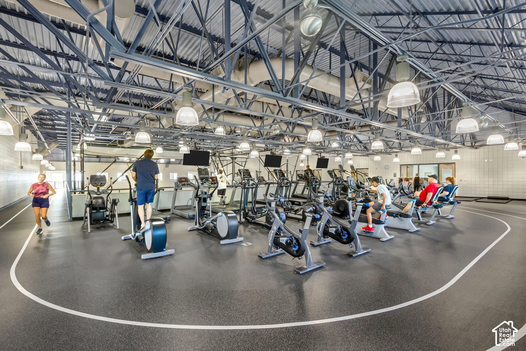 Industrial-style fitness center featuring an exposed truss ceiling, resilient rubber flooring, an integrated running track, and multiple hanging pendant lights